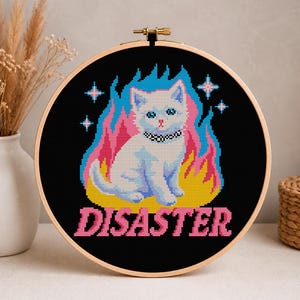 May include: A cross-stitch embroidery hoop featuring a white cat with blue eyes, a black collar, and a pink nose, set against a black background with flames in shades of pink, yellow, and blue. The word "DISASTER" is stitched in pink.
