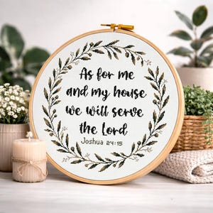 Pode incluir: Um bastidor de bordado em ponto cruz com o texto "As for me and my house we will serve the Lord Joshua 24:15". O design apresenta uma coroa de flores em tons de verde e castanho, sobre um fundo branco.