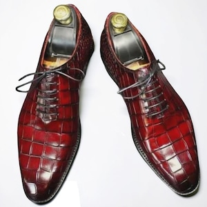 Peut inclure: Une paire de chaussures richelieu en cuir rouge foncé brillant avec un motif texturé en peau de crocodile. Chaque chaussure comprend des lacets noirs fins et un embauchoir en plastique noir avec un bouton doré visible au talon.