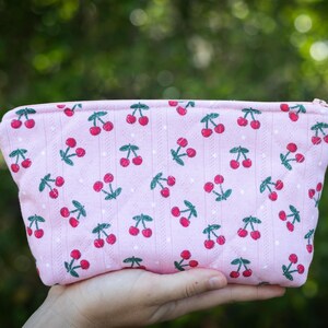 May include: A pink zippered pouch with a repeating pattern of red cherries and green leaves. The pouch is held in a hand against a blurred green background. The cherries are evenly spaced across the fabric.