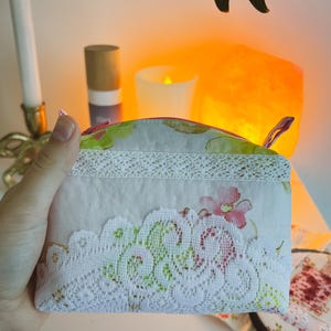 Peut inclure: Petite pochette rectangulaire à imprimé floral avec une bordure en dentelle blanche. La pochette est dotée d'une fermeture éclair avec une tirette rose. Le motif floral comprend des fleurs roses et vertes sur fond clair. La pochette est tenue par une main.