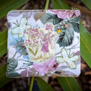 May include: A square coin purse with a floral pattern and a zip. It features appliqued butterflies and a fairy design. The background has pink, green, and white floral patterns. Small decorative pins and a yellow flower accent the design.