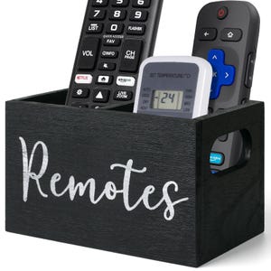 May include: A black wooden box with the word "Remotes" in white script. The box holds several remote controls and a small digital thermostat. The remotes are black and gray, with various buttons and labels.