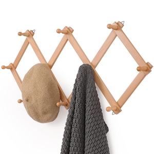 May include: A light-colored wooden accordion-style wall rack with multiple pegs. A tan beret and a gray scarf hang from the pegs. The rack is mounted on a white wall.