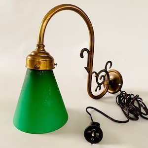 Könnte beinhalten: Eine Vintage-Wandleuchte mit Messing-Finish und grünem Glasschirm. Die Lampe hat einen gebogenen Arm und eine dekorative Rückplatte. Das Netzkabel und der Stecker sind sichtbar. Die Lampe ist für den Innenbereich konzipiert.