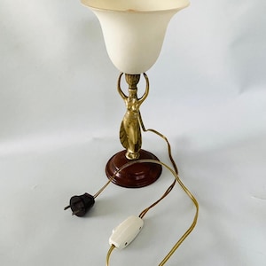May include: Vintage table lamp featuring a cream-colored, tulip-shaped shade. The lamp is supported by a brass figure and a dark brown wooden base. It has a cord with a switch and a plug.