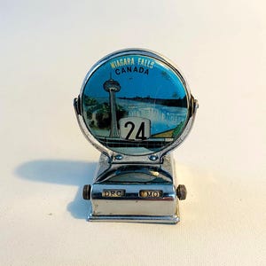 Op de afbeelding: Een vintage metalen bureaukalender met een ontwerp van de Niagara Falls, Canada. De kalender toont het nummer 24 op een blauwe achtergrond met een afbeelding van de watervallen en een toren. De basis heeft draaiknoppen voor de maand en de dag.