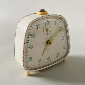 Könnte beinhalten: Ein cremefarbener Peter-Wecker im Vintage-Stil mit abgerundeter quadratischer Form. Die Uhr hat grüne Ziffern, ein kleines Zifferblatt und gelbe Zeiger. Die Uhr hat einen kleinen gelben Knopf oben und steht auf vier kleinen gelben Füßen.