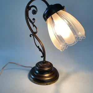 Könnte beinhalten: Antike Tischlampe mit bronzefarbenem Sockel und geschwungenem Stiel. Die Lampe hat einen Lampenschirm aus mattiertem Glas in Blütenform, der ein weiches Licht ausstrahlt. Das Design umfasst dekorative Schnörkel und eine dunkelbronze Oberfläche. Höhe: ca. 30 cm.