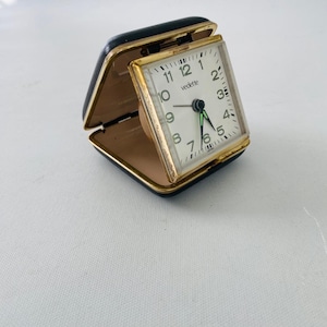 Op de afbeelding: Een vintage Vedette reiswekker. De klok heeft een vierkante wijzerplaat met zwarte cijfers en wijzers, gezet in een goudkleurig frame. De klok is verpakt in een opvouwbare zwarte kunstlederen hoes. De wijzerplaat is ongeveer 5 cm breed.