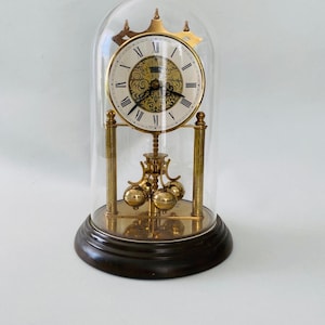 May include: A vintage brass clock with a white face and black Roman numerals, enclosed in a glass dome. The clock has a pendulum with three brass spheres and rests on a dark wooden base. The clock's hands are black.