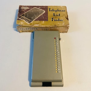 Op de afbeelding: Een vintage telefoonlijstzoeker met een grijze plastic behuizing en een kartonnen doos. De doos is voorzien van het opschrift "Telephone List Finder" en toont een diagram van het apparaat. De zoeker heeft gelabelde tabbladen.
