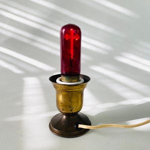 May include: A small brass lamp base with a cylindrical red glass bulb featuring a cross-shaped filament inside. The lamp has a flared top, a circular base, and a light-coloured electrical cord extending from the side of the metal housing.