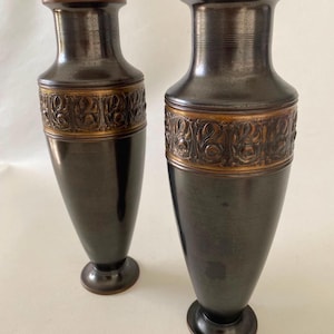 Könnte beinhalten: Zwei dunkelmetallische Vasen mit glatter Oberfläche und einem breiten Mittelband, das ein erhabenes, verziertes Schnörkelmuster in Bronzeton aufweist. Jede Vase hat einen schmalen Hals, einen ausgestellten Rand und einen runden Fuß.