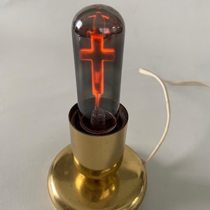 May include: A table lamp with a polished gold-tone metal base and a cylindrical glass bulb. The bulb features a glowing orange neon filament shaped like a cross inside a dark, smoky-tinted glass casing, connected to a white electrical cord.