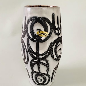 Könnte beinhalten: Eine zylindrische Keramikvase mit weiß glasierter Oberfläche und einem handgemalten schwarzen abstrakten Muster aus kreisförmigen und linearen Motiven. Ein goldener ovaler Aufkleber mit dem schwarzen Schriftzug Jasba klebt vorne.