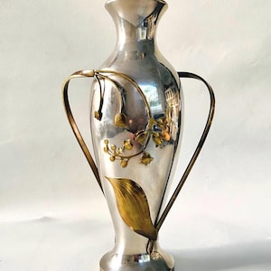 Pode incluir: Um vaso de metal prateado alto com pescoço estreito e borda alargada. O corpo é adornado com motivos florais e de folhas aplicados na cor latão. Duas alças finas e curvas estendem-se do pescoço até à parte inferior do corpo do vaso.