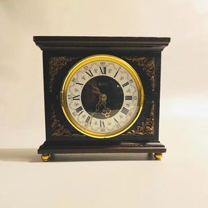 May include: A dark brown wooden mantel clock with a gold-coloured clock face and Roman numerals. The clock has decorative gold accents on the corners and base, and gold-coloured feet. The clock face has the word "JEKA" printed on it.