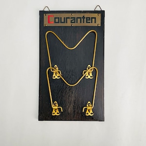 Pode incluir: Um cabide de parede vintage com base de madeira preta e detalhes em metal dourado. A palavra "Couranten" é exibida em letras vermelhas e douradas no topo. O cabide apresenta um design exclusivo com uma barra central curva e quatro ganchos.
