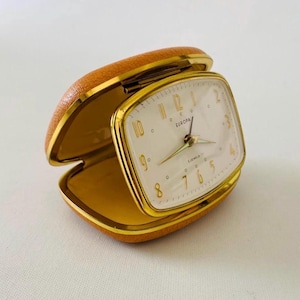Op de afbeelding: Een vintage Europa reiswekker met een beige, gestructureerde, lederachtige opvouwbare behuizing en een goudkleurig metalen frame. De wijzerplaat is wit met goudkleurige cijfers, wijzers en de tekst Europa 2 Jewels in het midden.