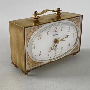 May include: A vintage gold-toned Jazitor electric travel clock with an oval face. The clock features a handle, decorative trim, and gold hands and numerals against a white background. The clock is rectangular with small feet.