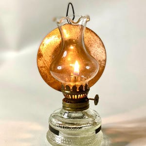 Può includere: Una piccola lampada a olio in stile antico con un serbatoio in vetro e un bruciatore color ottone. La lampada ha una base in vetro trasparente con un design strutturato e un riflettore color rame. All'interno è accesa una piccola fiamma.