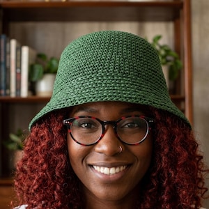 May include: A green crochet bucket hat with a wide brim. The hat is made of a textured yarn and has a slightly sloped crown. The hat is worn by a person with red curly hair and glasses.