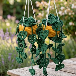Puede incluir: Dos macetas colgantes de ganchillo con vides de hojas verdes. Las macetas son amarillas y están suspendidas por cuerdas blancas. Las hojas son de color verde oscuro y tienen un diseño en forma de corazón. El fondo está borroso, con toques de vegetación y flores.