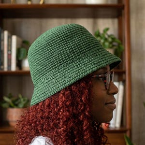 May include: A green crochet bucket hat. The hat is made of a textured yarn and has a slightly sloped brim. The hat is worn by a person with red curly hair and glasses.