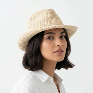 May include: A beige fedora-style hat with a woven texture. The hat has a structured crown and a wide brim. The hat is worn by a person wearing a white collared shirt. The background is plain white.