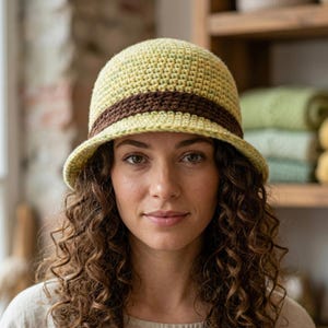 Könnte beinhalten: Gehäkelter Glockenhut in Hellgrün mit braunem Band. Der Hut hat eine runde Krone und eine kleine Krempe. Der Hut ist handgefertigt und scheint aus Garn zu bestehen.