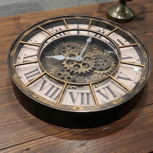 Op de afbeelding: Een ronde wandklok in antieke stijl met een zwart frame en een crèmekleurige wijzerplaat met Romeinse cijfers. De klok heeft goudkleurige wijzers en een zichtbaar tandradmechanisme. De klok heeft een diameter van ongeveer 30 cm.