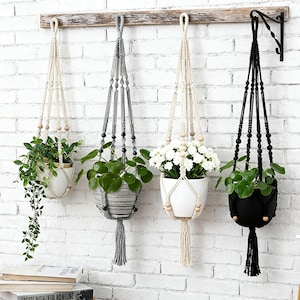 Może przedstawiać: Cztery makramowe wieszaki na rośliny z doniczkami i roślinami na tle białej ceglanej ściany. Wieszaki są kremowe, szare i czarne, z drewnianymi koralikami i frędzlami. Rośliny obejmują zielone rośliny liściaste i białe kwiaty.