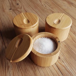 Puede incluir: Tres saleros de bambú con tapas y cucharas pequeñas a juego. Un salero está abierto, revelando un montón de sal blanca. Los saleros son cilíndricos y están sobre una superficie de madera.