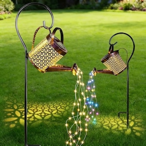 Puede incluir: Dos regaderas decorativas con energía solar, con acabado bronce y diseño calado, se exhiben en un jardín. Están suspendidas de estacas de metal negro. Cadenas de luces LED blancas cálidas y de colores caen de las boquillas.