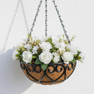 May include: A hanging basket filled with white roses and greenery. The basket has a decorative black metal frame with heart-shaped designs and a brown liner. The basket is suspended by a black chain against a white wall.