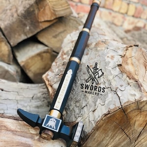May include: A black and gold hammer with a dark wooden handle, displayed on a pile of wood. The hammer has a square head and a decorative emblem. The text "SWORDS - MAKERS" is visible.