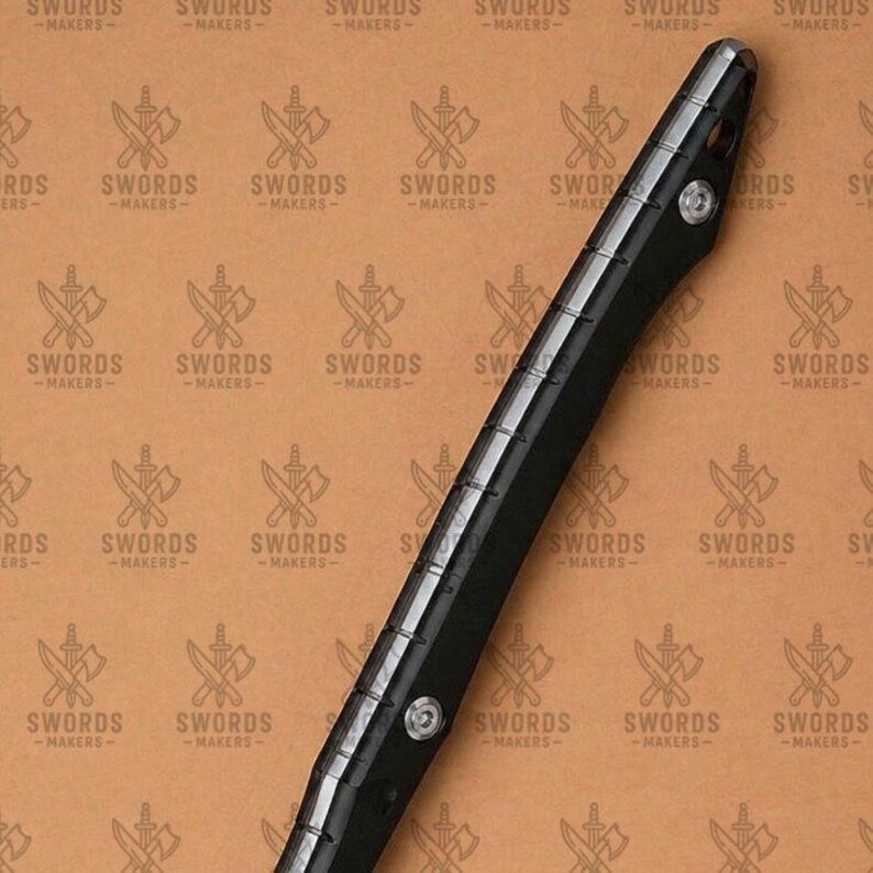 May include: A black sword handle with silver accents and rivets. The handle is long and slender, with a textured grip. The background features a repeating pattern of crossed swords and axes with the text "SWORDS -MAKERS-".