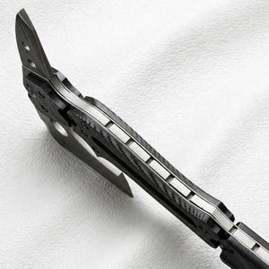 May include: Close-up of a tactical tomahawk with a black and silver design. The axe head features a sharp blade and a pointed spike. The handle has a textured, carbon fiber appearance.