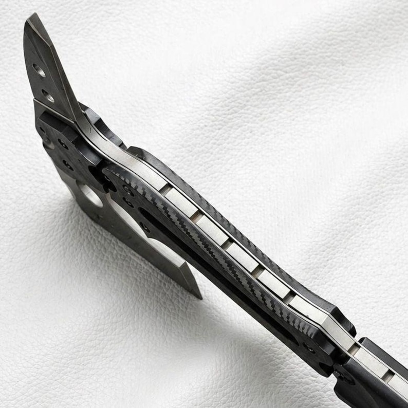 May include: Close-up of a tactical tomahawk with a black and silver design. The axe head features a sharp blade and a pointed spike. The handle has a textured, carbon fiber appearance.