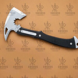 May include: A silver and black tactical tomahawk with a curved blade and a spiked end. The tomahawk has a black handle with a silver accent and a hole at the end. The text "SWORDS - MAKERS" is in the background.
