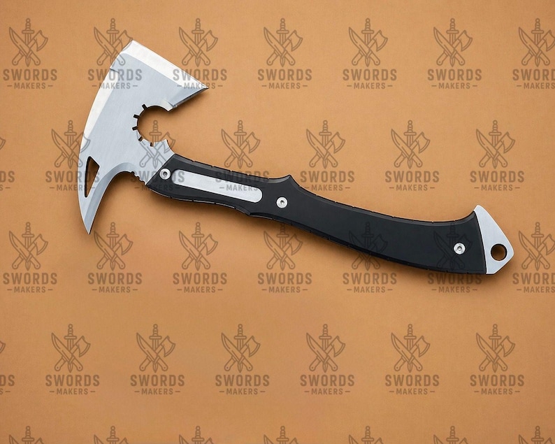 May include: A silver and black tactical tomahawk with a curved blade and a spiked end. The tomahawk has a black handle with a silver accent and a hole at the end. The text "SWORDS - MAKERS" is in the background.