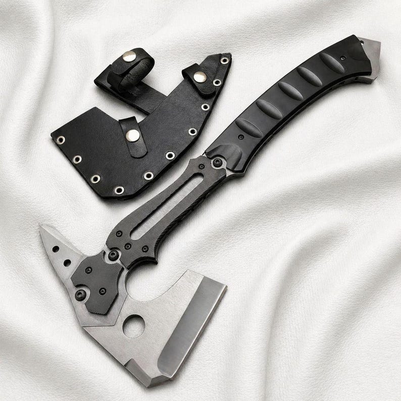 May include: A tactical axe with a black handle and a silver blade, accompanied by a black leather sheath. The axe features a unique design with a sharp edge and a pointed end. The sheath has a belt loop for easy carrying.
