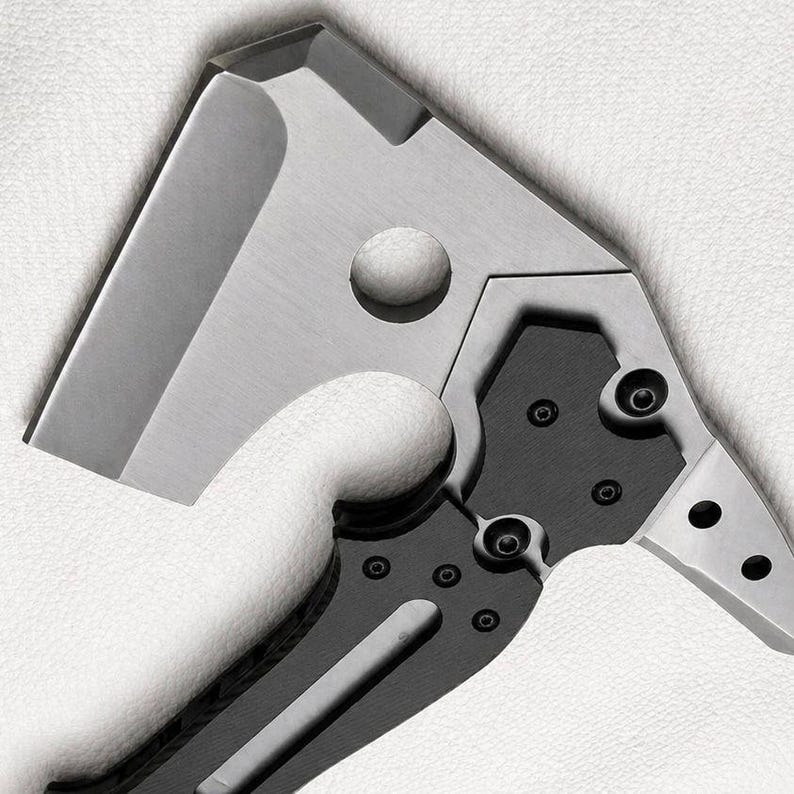 May include: Close-up of a stainless steel and black folding knife. The blade is a modified cleaver shape with a circular hole. The handle is black with silver accents and visible screws. The knife is designed for utility and everyday carry.