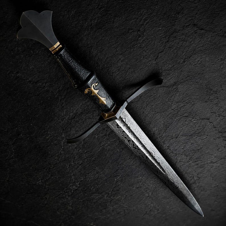 May include: A dagger featuring a patterned Damascus steel blade, a black textured grip, and a gold-coloured metal inlay. The hilt includes a black crossguard and a flared pommel, showcasing a sharp, pointed tip and intricate metalwork details.