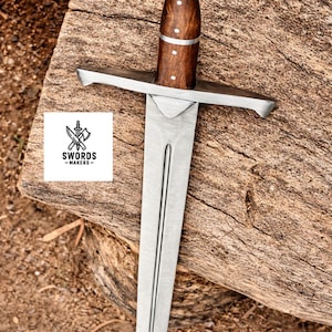 May include: A medieval-style sword with a polished steel blade and crossguard. The handle is crafted from dark wood, accented with small, white decorative studs. The pommel is a rounded, silver-colored metal. The sword rests on a weathered wooden surface. The logo "SWORDS -MAKERS-" is visible.