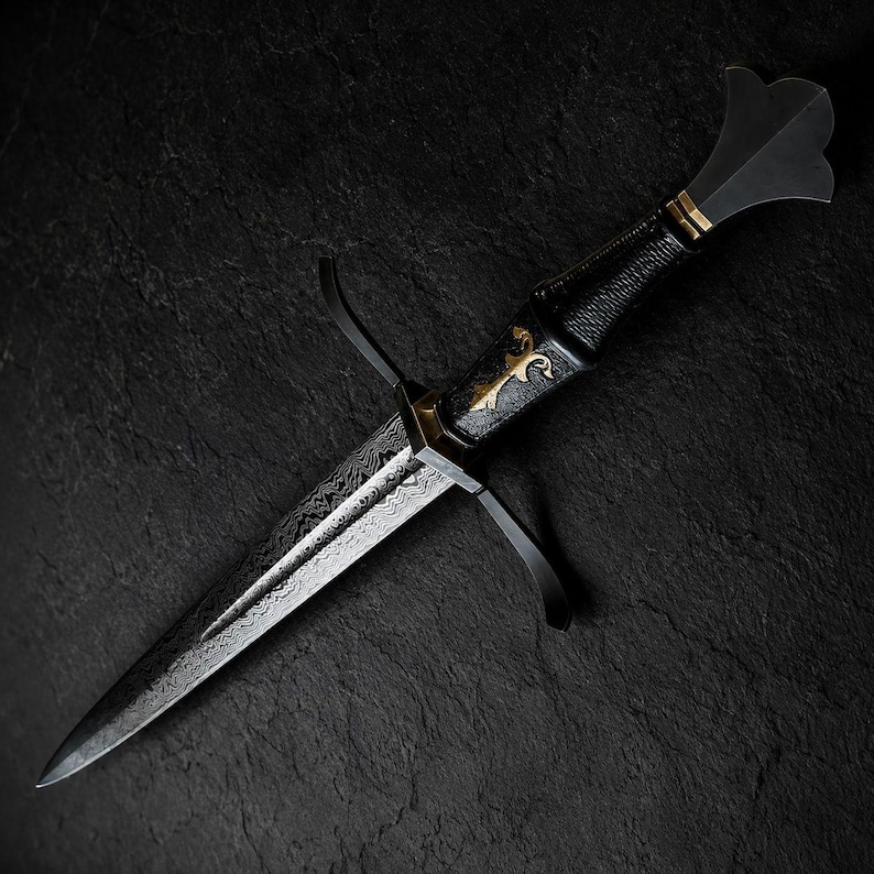 May include: A dagger featuring a patterned Damascus steel blade with a sharp point. The handle is black with a textured grip, accented by a gold-coloured inlay and a flared pommel. A black metal crossguard with curved quillons extends from the hilt.