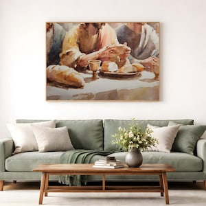 May include: Watercolor painting of a table scene with people, bread, and a cup. The artwork is framed and hangs above a green sofa with pillows, a wooden coffee table, and a potted plant. The painting has warm tones.