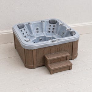 May include: A square hot tub featuring a light grey interior basin with multiple integrated water jets and dark grey headrests. The exterior is finished in a brown wood-look panelling, complemented by a matching two-step staircase attached to the front.