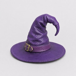 Peut inclure: Un chapeau pointu violet avec un large bord et une surface texturée. Le chapeau présente une bande violette assortie autour de la base, agrémentée d'un petit détail de boucle de couleur métallique. Le sommet est légèrement courbé et affaissé.